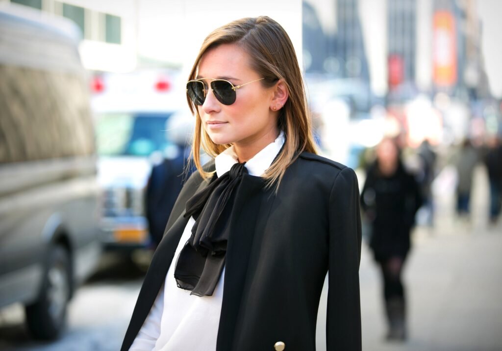 A woman wearing a sophisticated black bow tie blouse and dark sunglasses, exuding timeless elegance in a city setting.