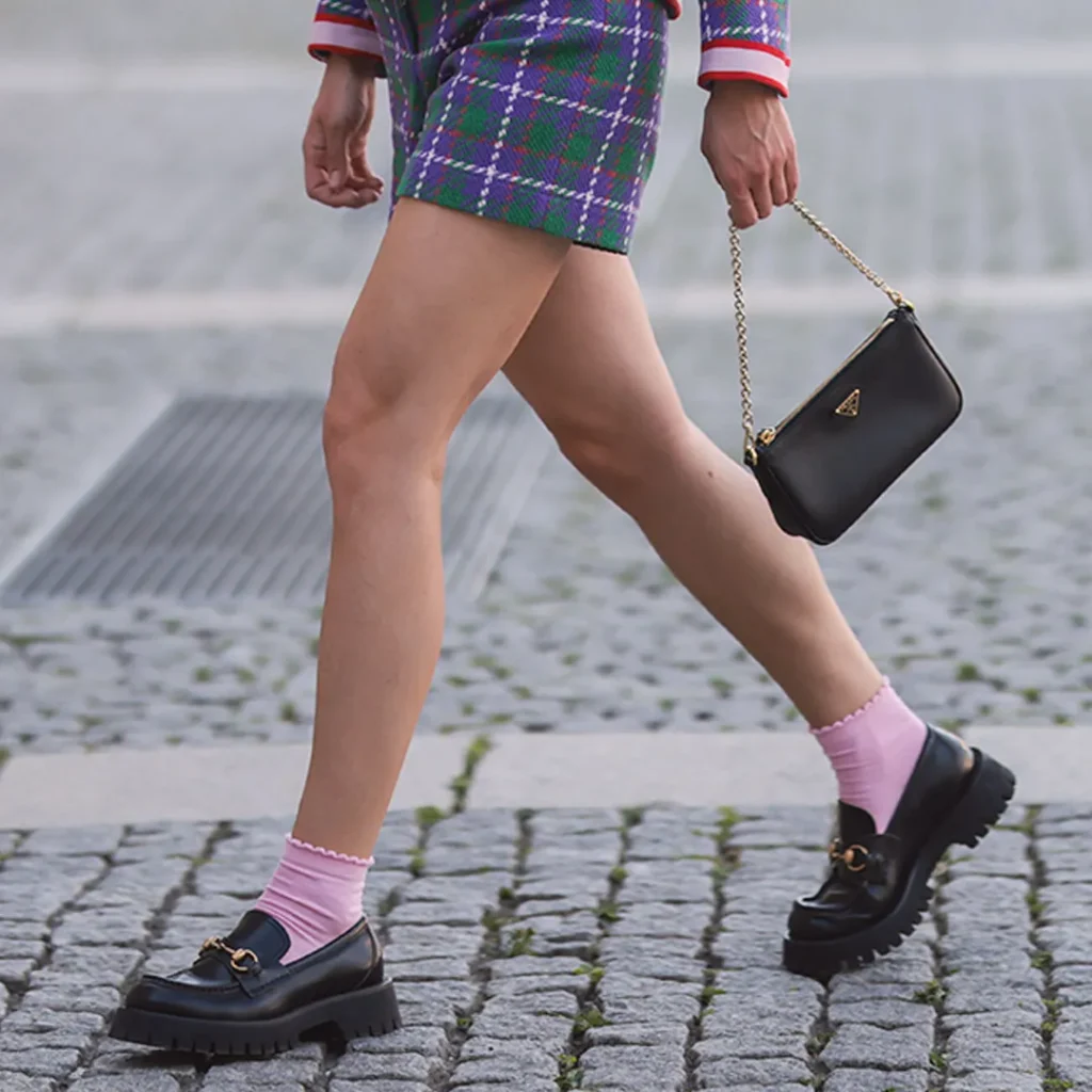 Close-up of a female wearing black chunky loafers with pink socks and a plaid mini skirt, walking on a cobblestone street.