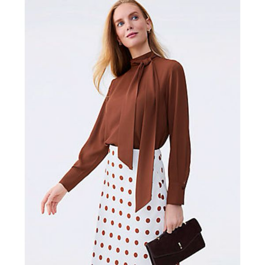 A woman wearing a chocolate brown Ann Taylor silk bow tie blouse paired with a white and black polka dot skirt, featured on The Posh EdiT.