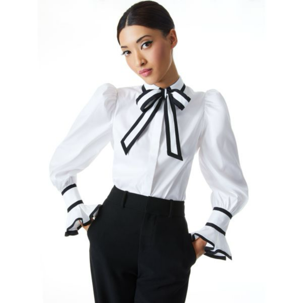 A woman wearing the Alice + Olivia Haren puff sleeve bow tie blouse in white with a black ribbon, styled with black trousers for a professional and sophisticated look on The Posh EdiT.