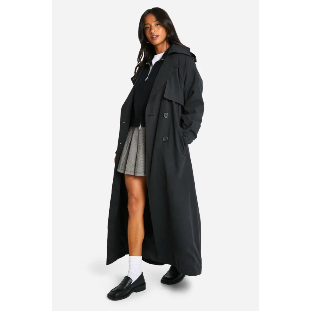 A woman wearing an oversized black double-breasted trench coat with a relaxed fit, styled over a grey skirt and white socks with black loafers.