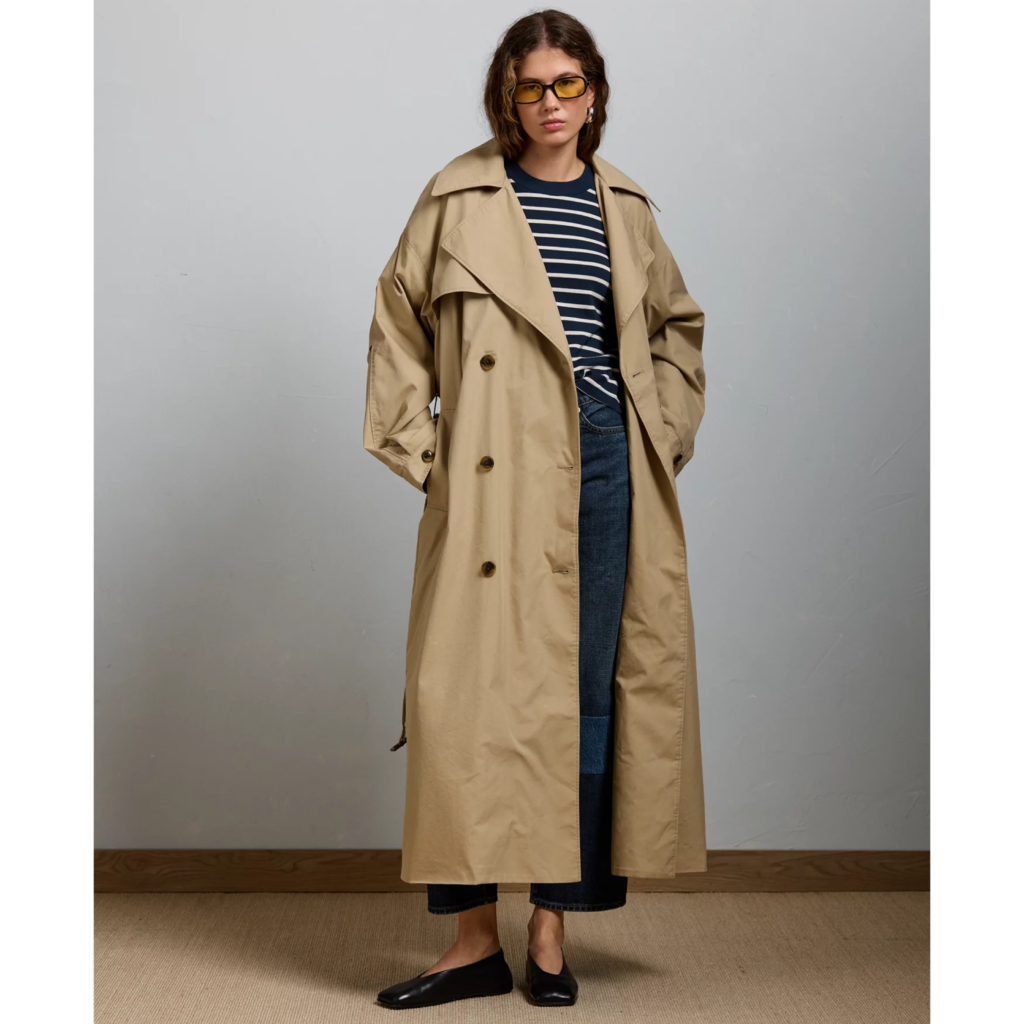 A woman wearing an oversized, long beige trench coat with a relaxed silhouette, styled over a striped navy top and dark midi skirt.