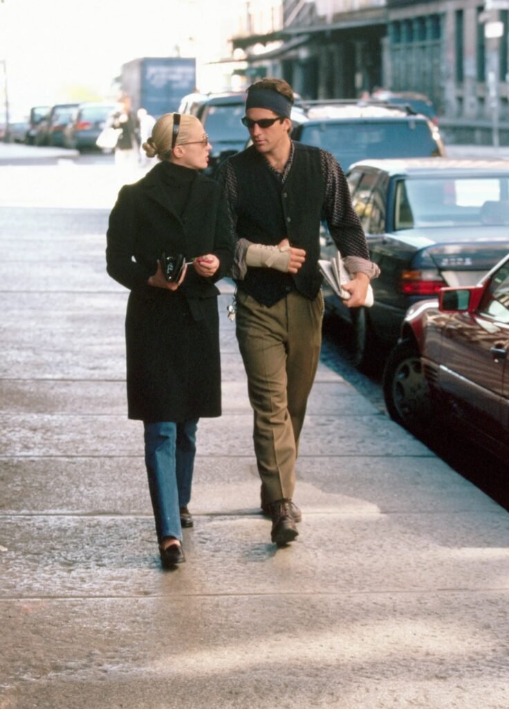 A candid street style photograph of Carolyn Bessette-Kennedy and John F. Kennedy Jr. walking in New York City; Carolyn wears a long black tailored coat and sunglasses.