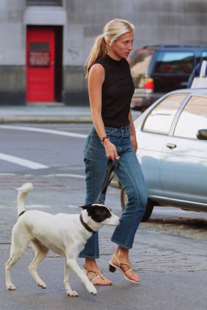 Carolyn Bessette-Kennedy walking her dog on a city sidewalk, wearing a black sleeveless mock-neck top, flare-leg blue jeans, and a simple black headband.