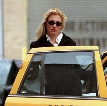 Carolyn Bessette-Kennedy leaning into a yellow New York City taxi cab, wearing a black blazer, white collared shirt, and her signature slim oval sunglasses.