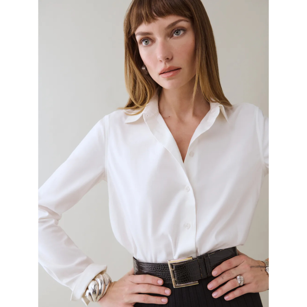 A model wearing the Reformation Alfie Relaxed Shirt in white silk, tucked into a black belt with a gold buckle, styled with layered gold bracelets for a refined 90s minimalist look